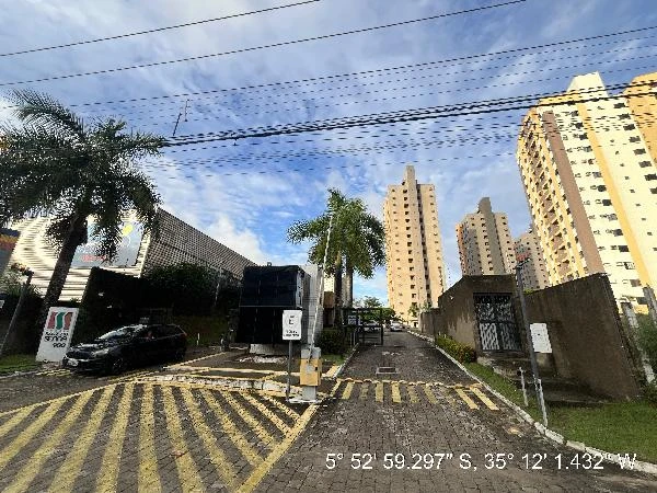 Apartamento em condomínio com blocos altos e guarita na entrada, área externa com árvores e calçadas pavimentadas sob céu nublado.