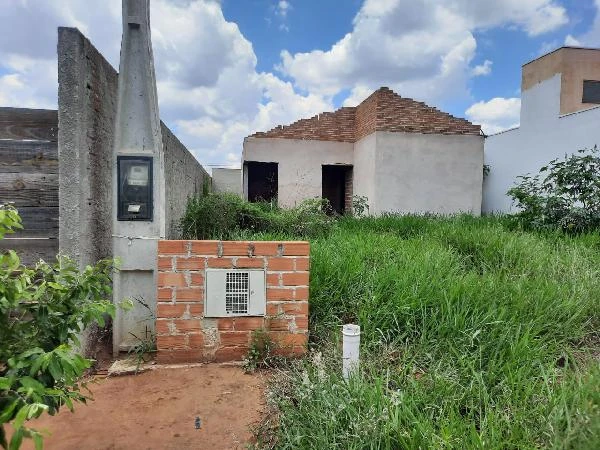 Casa em construção com estrutura de tijolos aparentes em terreno com vegetação alta e muro baixo ao redor