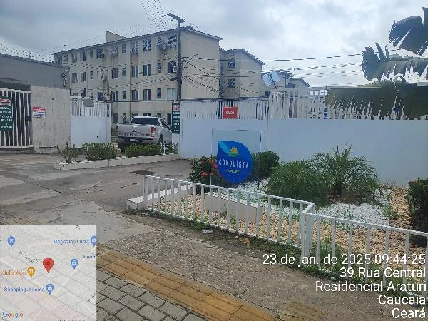 Apartamento em condomínio com portão branco, área externa com jardim e prédios residenciais ao fundo em Caucaia Ceará