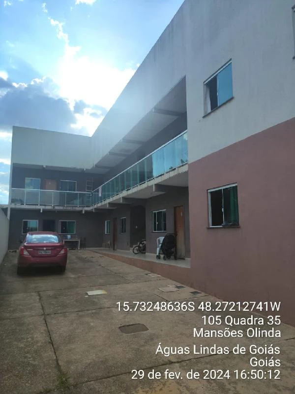 Apartamento cinza com sacada de vidro e vaga de garagem em pátio com carro vermelho e céu nublado