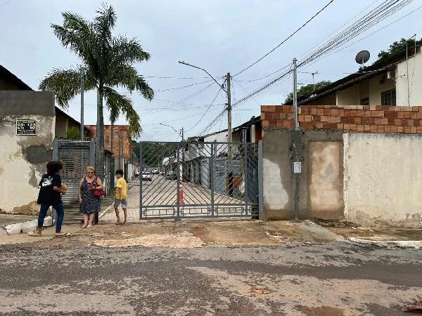 Condomínio residencial com portão de metal e muro de alvenaria em área urbana com árvores e pessoas na entrada