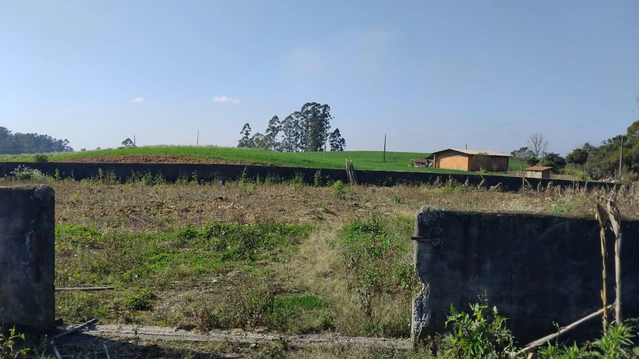 Terreno rural de 1600m² com vegetação rasteira e cerca baixa em área aberta e casas ao fundo