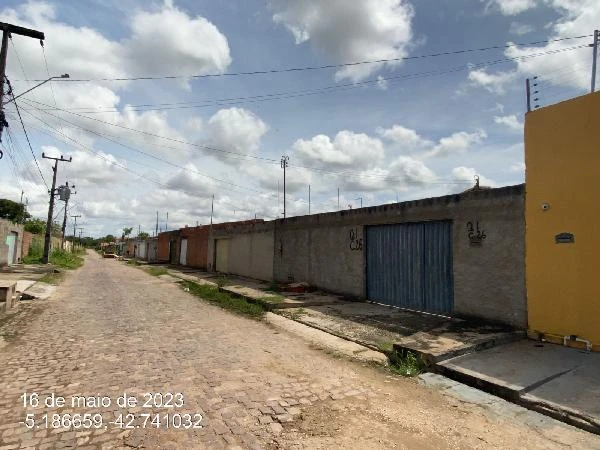 Casa com muro alto e portão metálico azul em rua de paralelepípedos com céu parcialmente nublado