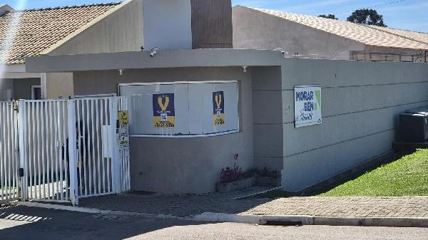 Casa cinza em condomínio com portão branco, fachada simples e muro baixo, sem avarias aparentes
