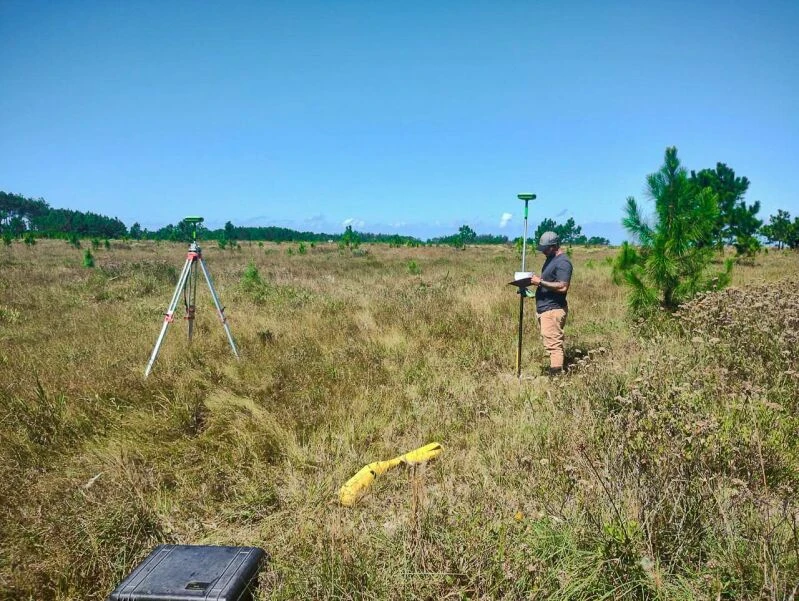 Topógrafo realizando a medição de um terreno amplo e plano com vegetação rasteira, utilizando equipamento de GPS em tripé e bastão, sob céu azul.
