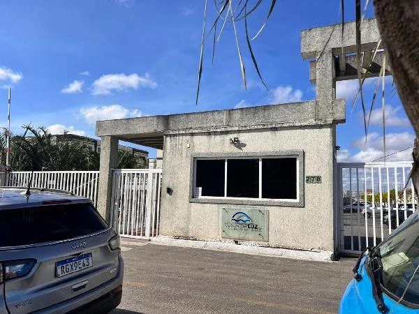 Entrada de condomínio residencial com portaria, muro cinza e carros estacionados em rua pavimentada