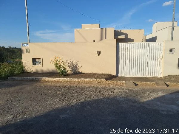 Casa térrea com parede bege, portão branco de metal e jardim pequeno em área urbana ensolarada