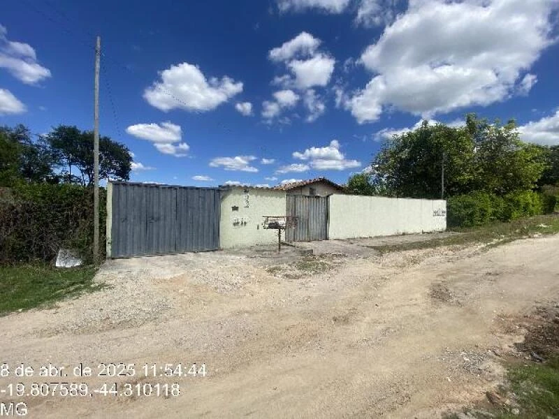 Casa com muro branco e portão metálico cinza em terreno com acesso de terra céu azul com nuvens
