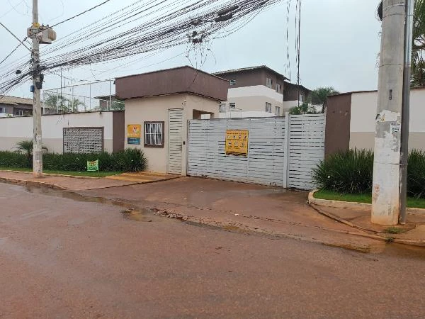 Apartamento em condomínio residencial com portão branco e muro alto, área externa com calçadas e vegetação próxima