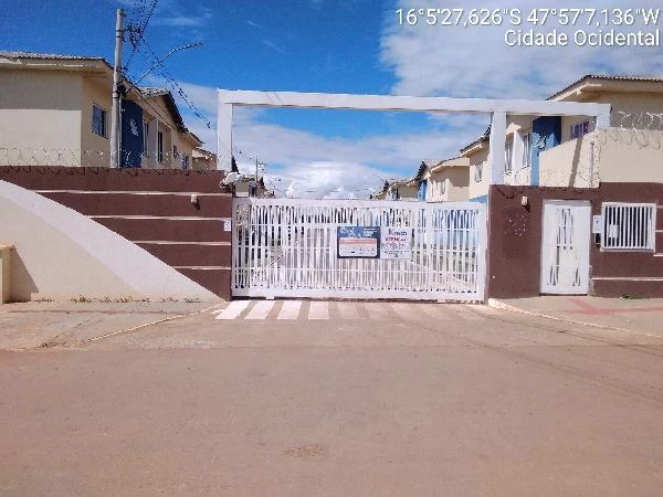 Apartamento em condomínio residencial com portão branco de metal e casas geminadas ao fundo em área urbana