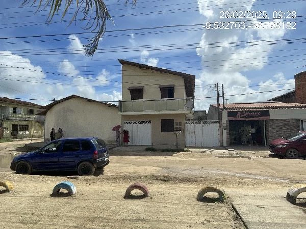 Casa branca de dois andares com varanda e portão branco em rua de terra, próximo a outros imóveis e carros estacionados