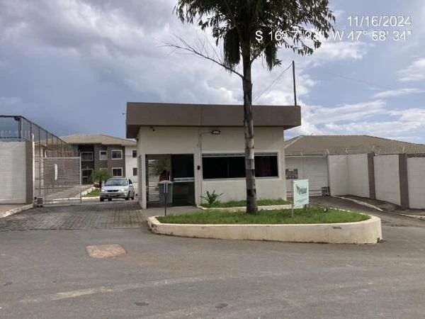 Apartamento em condomínio com guarita de segurança e carro estacionado na entrada, fachada em bom estado sob céu nublado