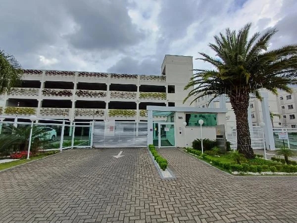 Apartamento branco com fachada de garagem coberta em condomínio com jardim e palmáceas ao redor sob céu nublado