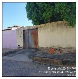 Casa com muro de concreto e portão de metal enferrujado em terreno plano vista externa com calçada e árvore