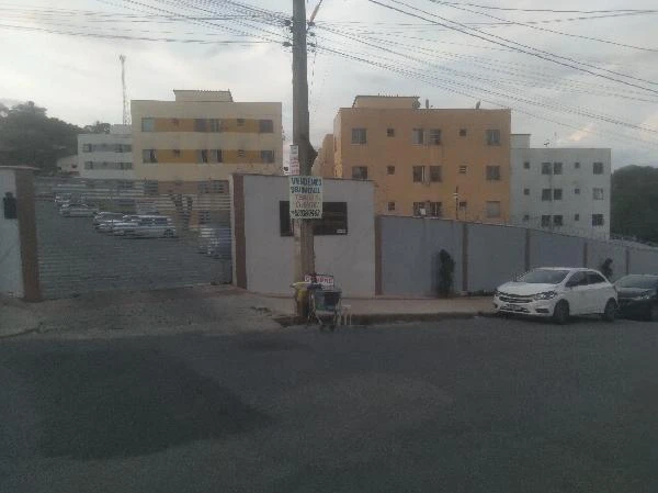 Apartamento branco em condomínio residencial com portão eletrônico e área de estacionamento visíveis na fachada