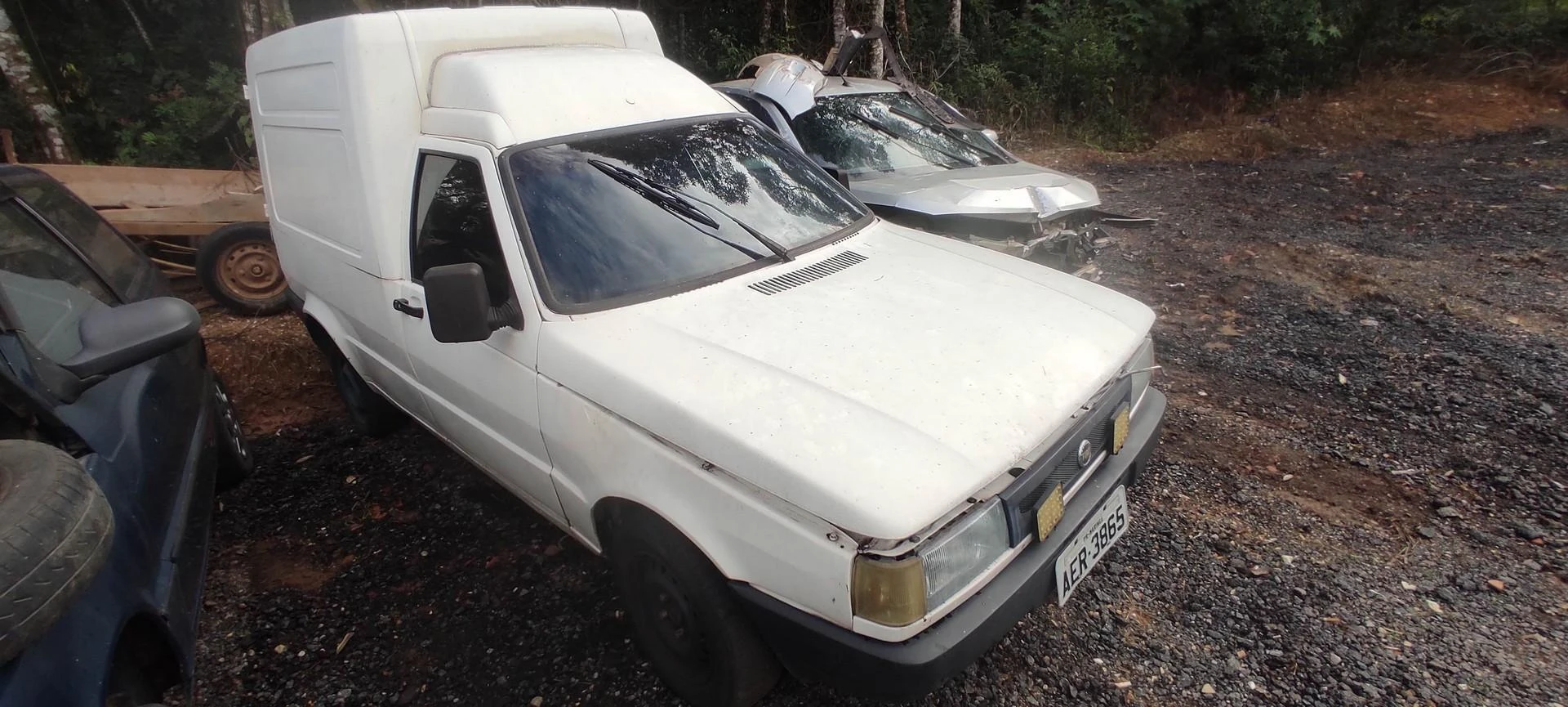 Fiat Fiorino branco 1989 em lote de veículos, com lataria desgastada e sinais de uso, estacionado em terreno de cascalho ao lado de outros carros danificados, indicando estado de sucata e necessidade de reparos ou reaproveitamento.